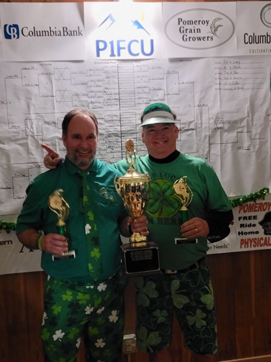 Two men in green, holding trophies, celebrating a win, in front of a tournament bracket.