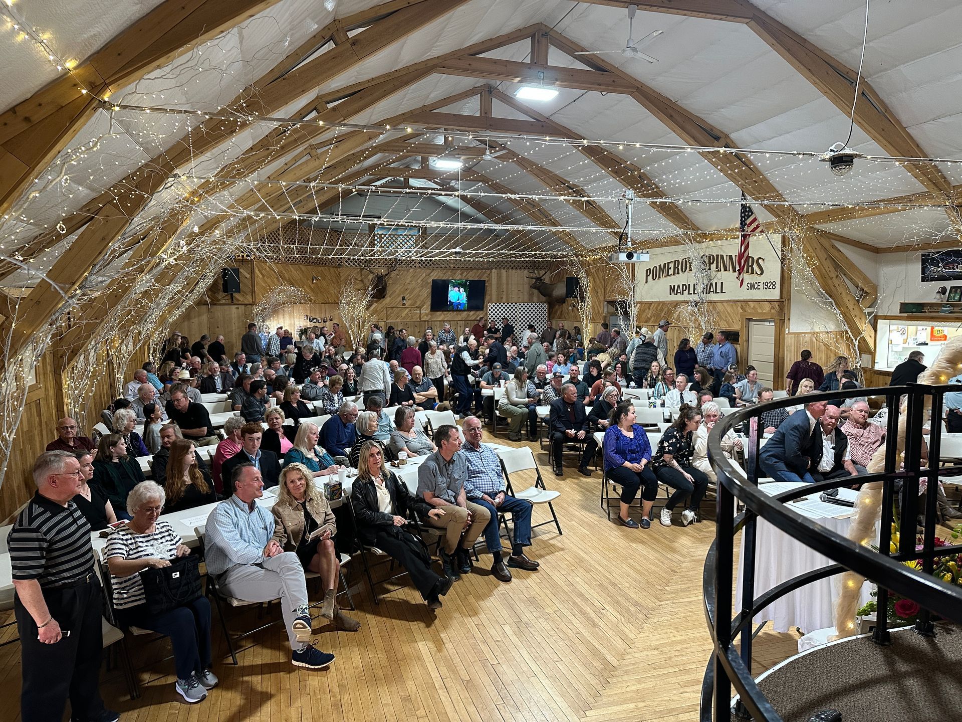 Large event in a wood-paneled hall with tables and chairs filled with people. String lights hang from the arched ceiling.