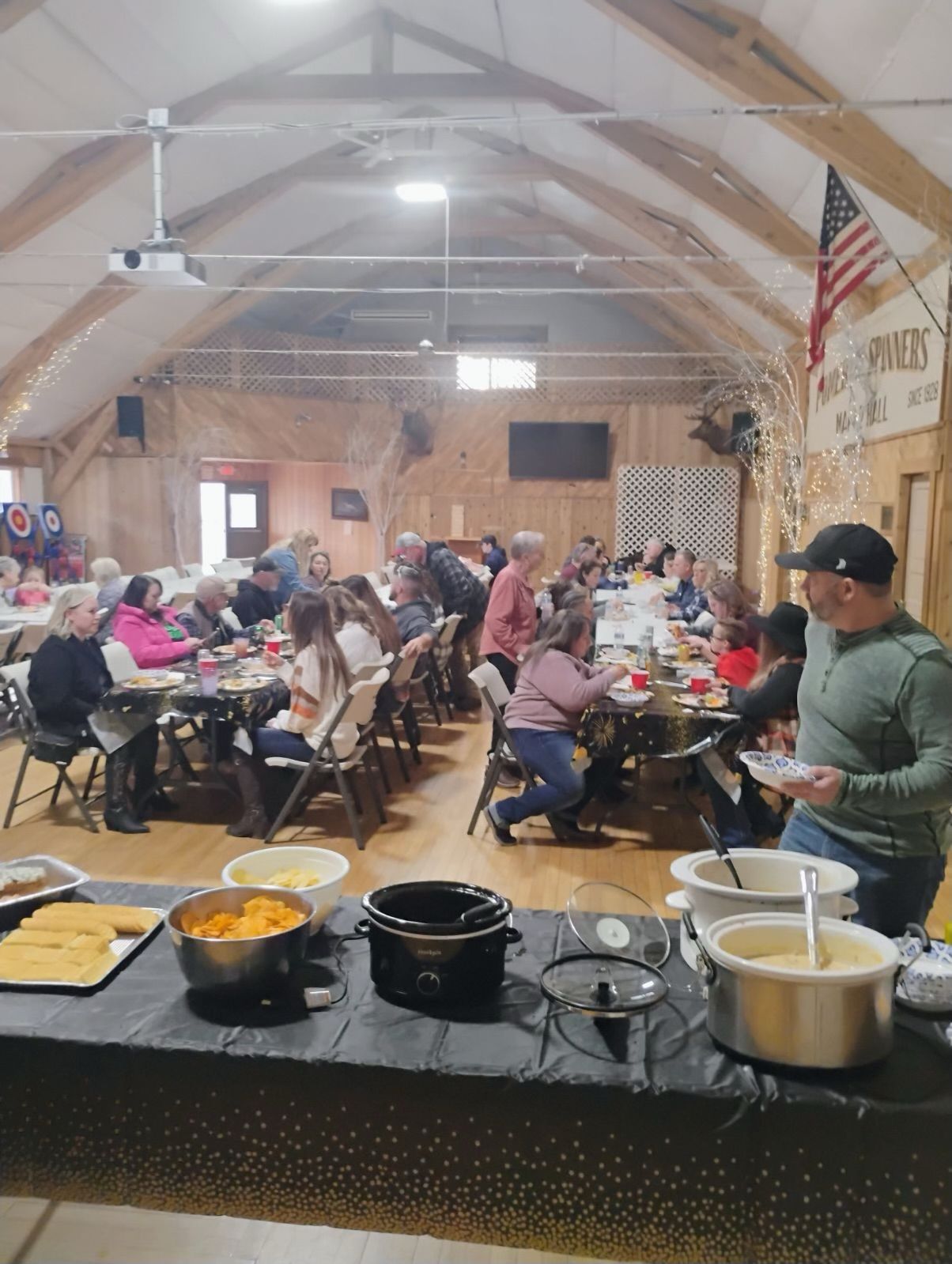 A community gathering in a hall with people eating at long tables. Food buffet in the foreground.