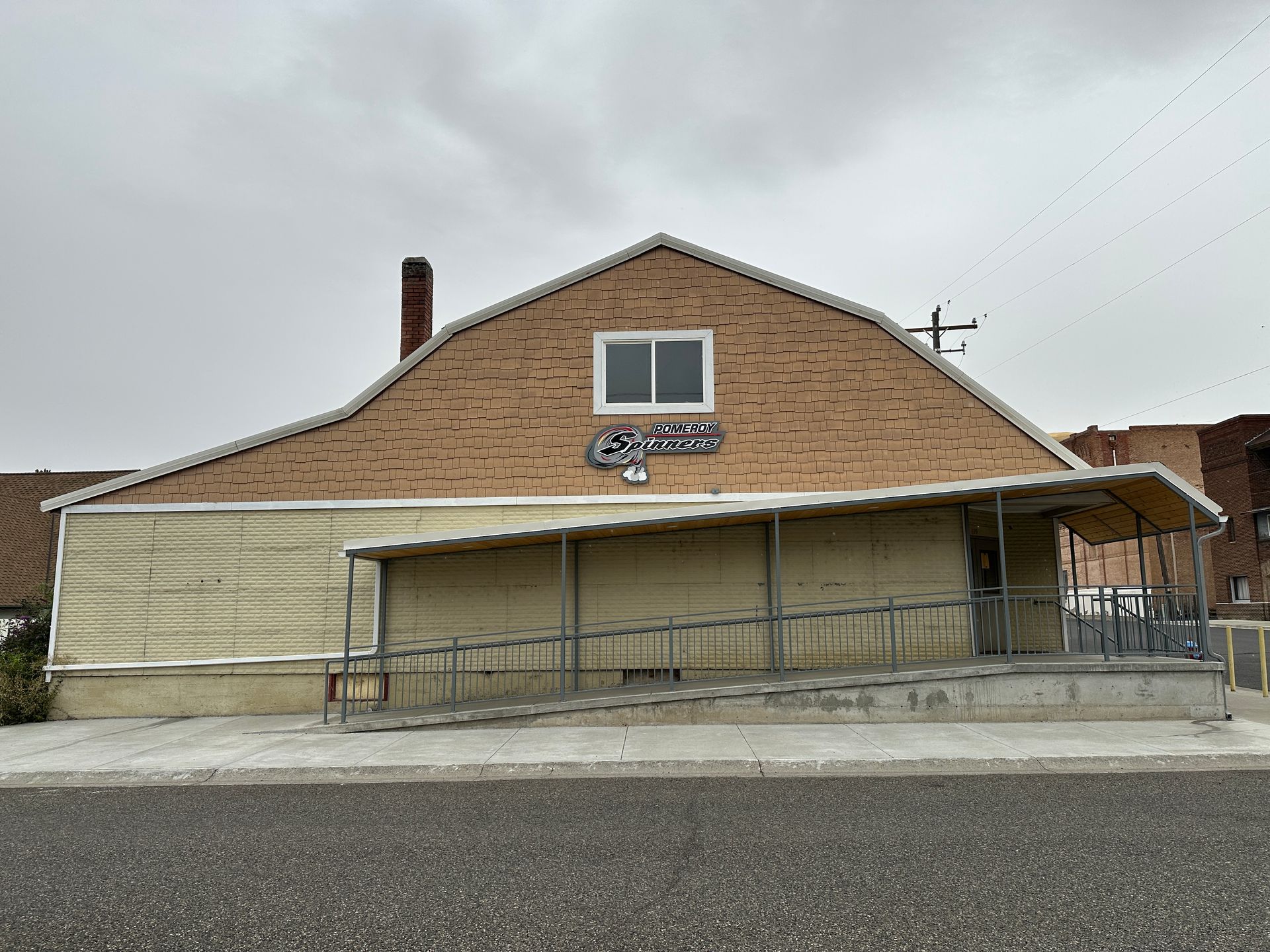Building with a ramp and awning, tan exterior, a sign above the entrance. Overcast sky.