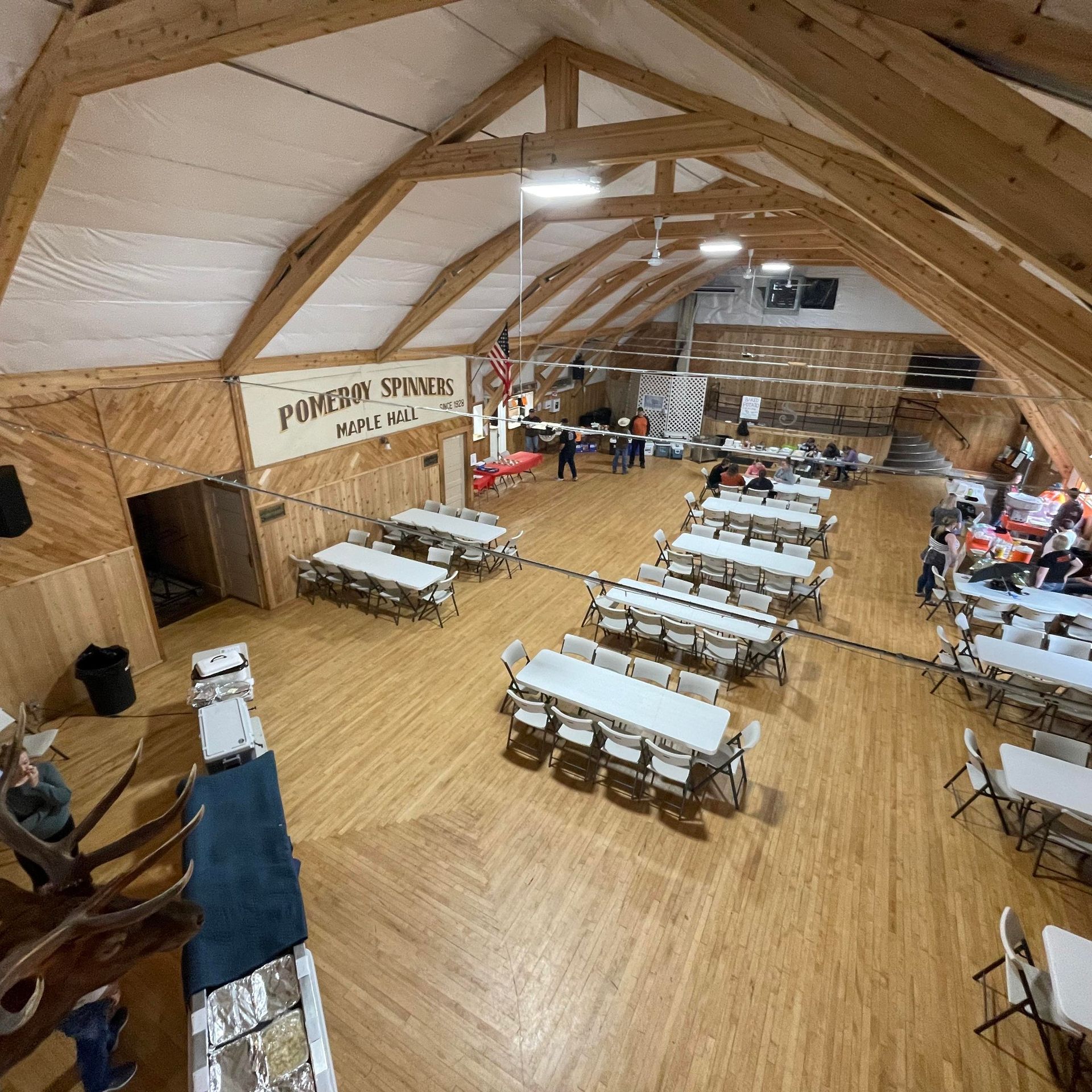 Large hall with wooden floors, tables set up, people milling about; 