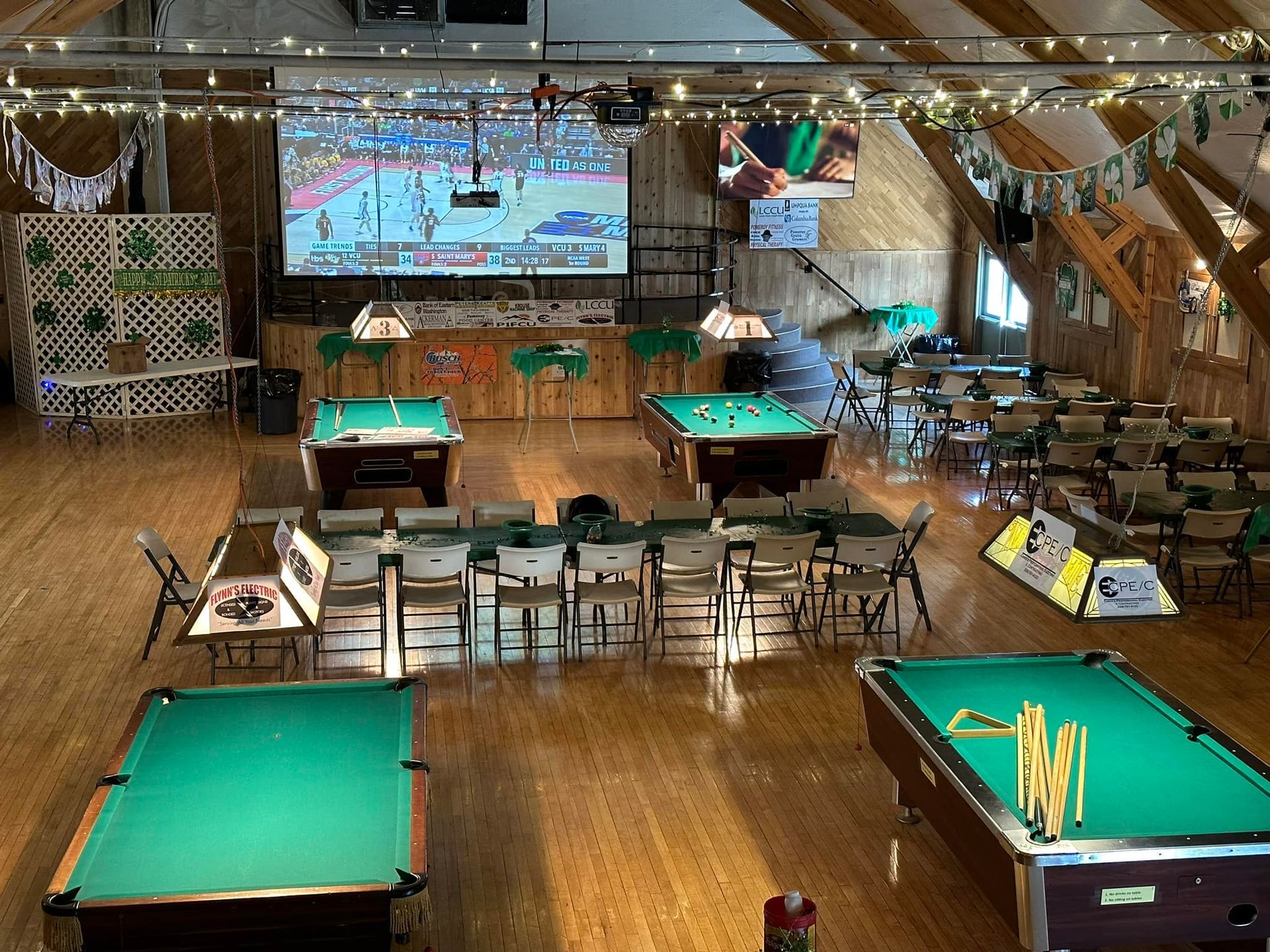An interior shot of a pool hall, with four pool tables, a projected screen, and seating.