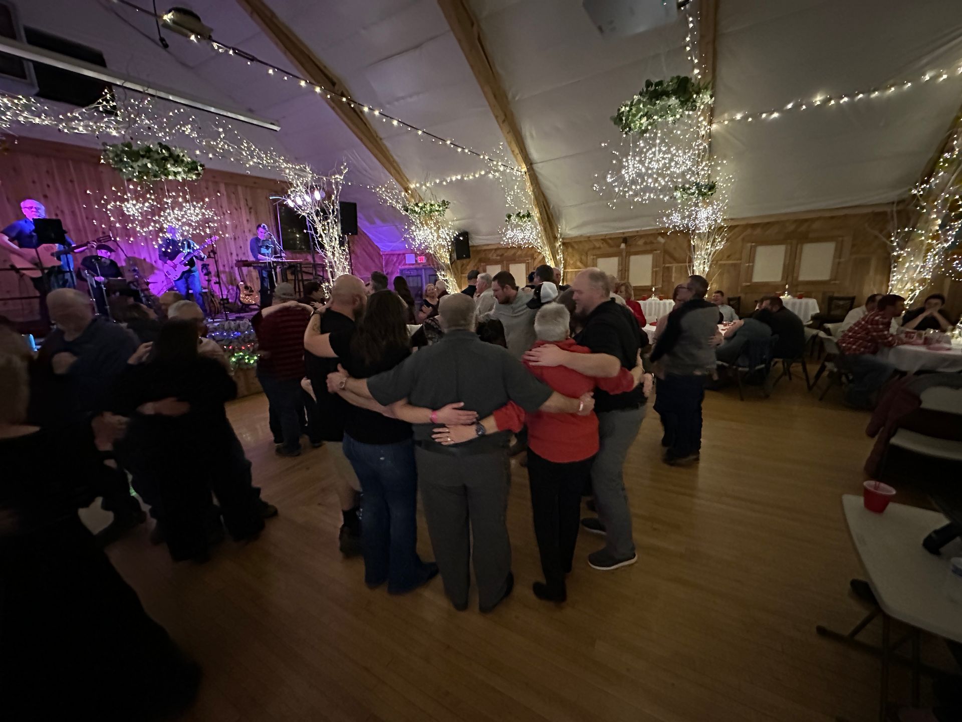 People dancing in a large hall with string lights and a band on stage.