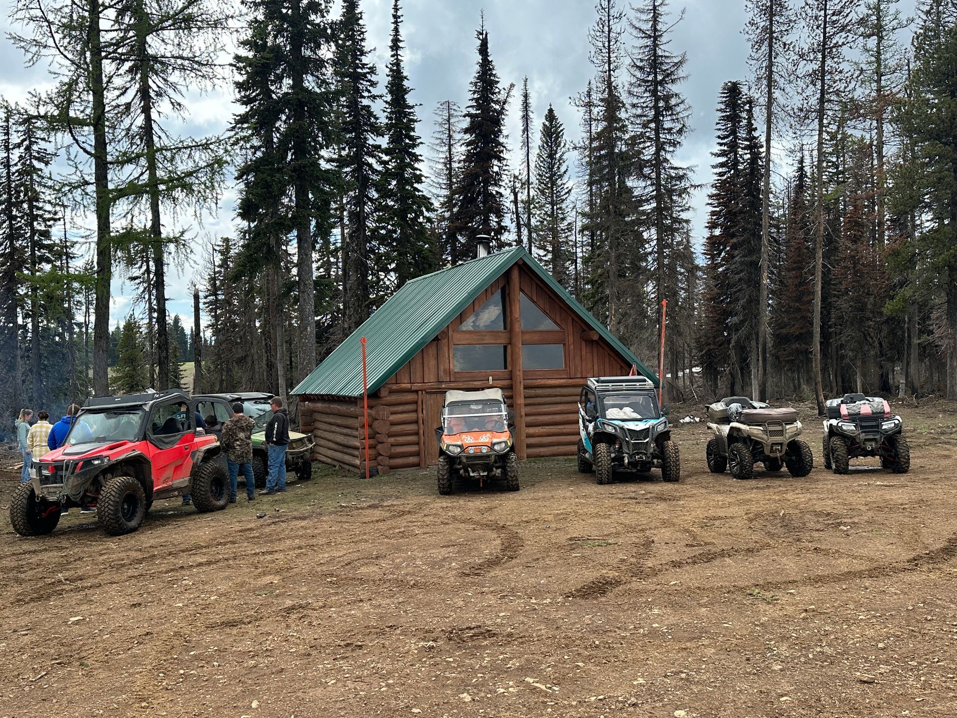 Off-road vehicles parked in front of a log cabin in a wooded area; people gathered nearby.