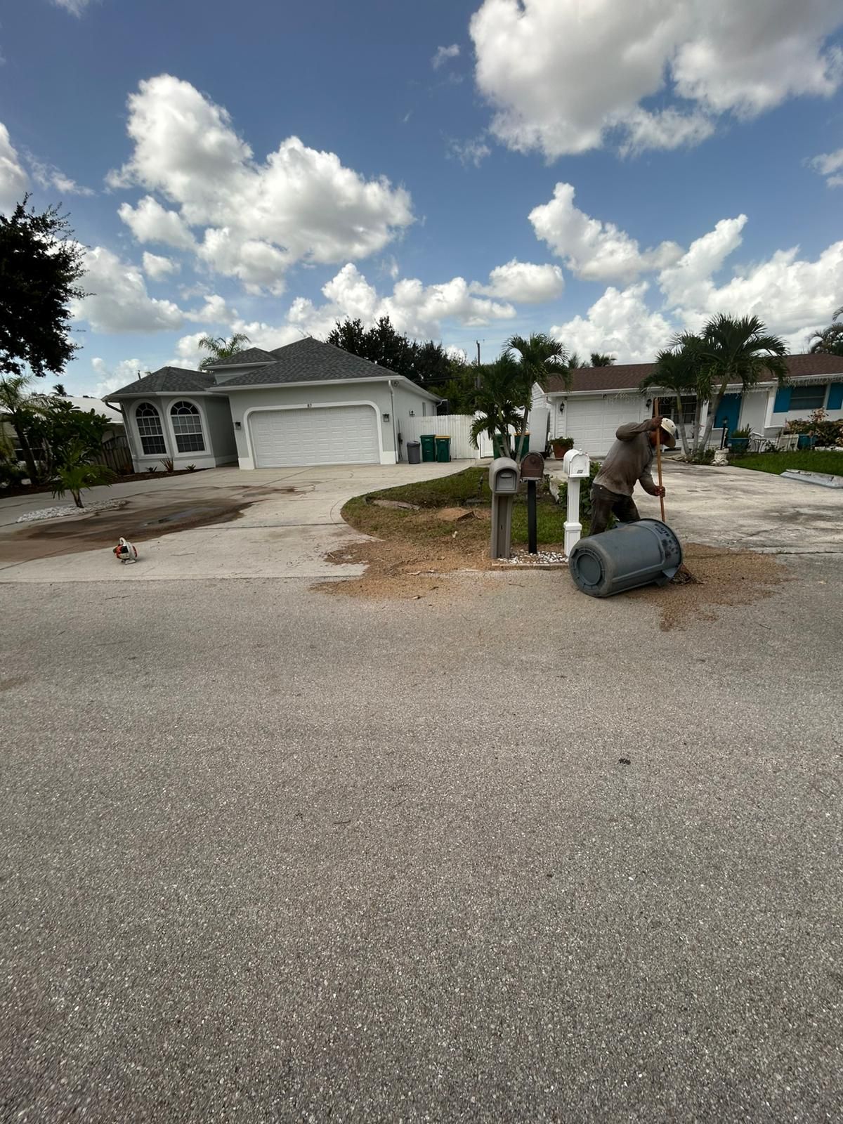 Man empties trash bin onto driveway in front of a house with blue sky and clouds.