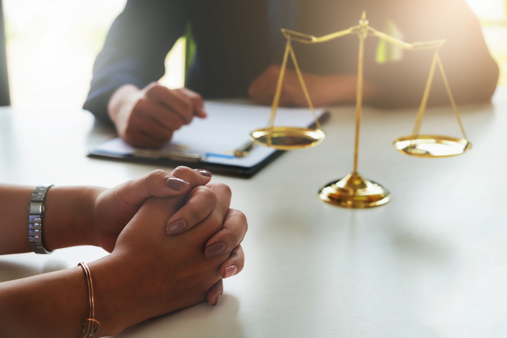 Hands clasped at a legal consultation, with a judge’s scales on a desk and a person writing in the background