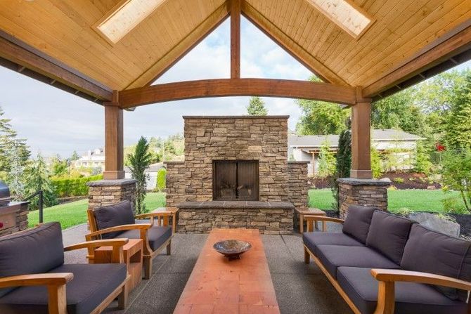 Covered outdoor patio with stone fireplace, wood beams, and dark cushioned seating overlooking a green yard