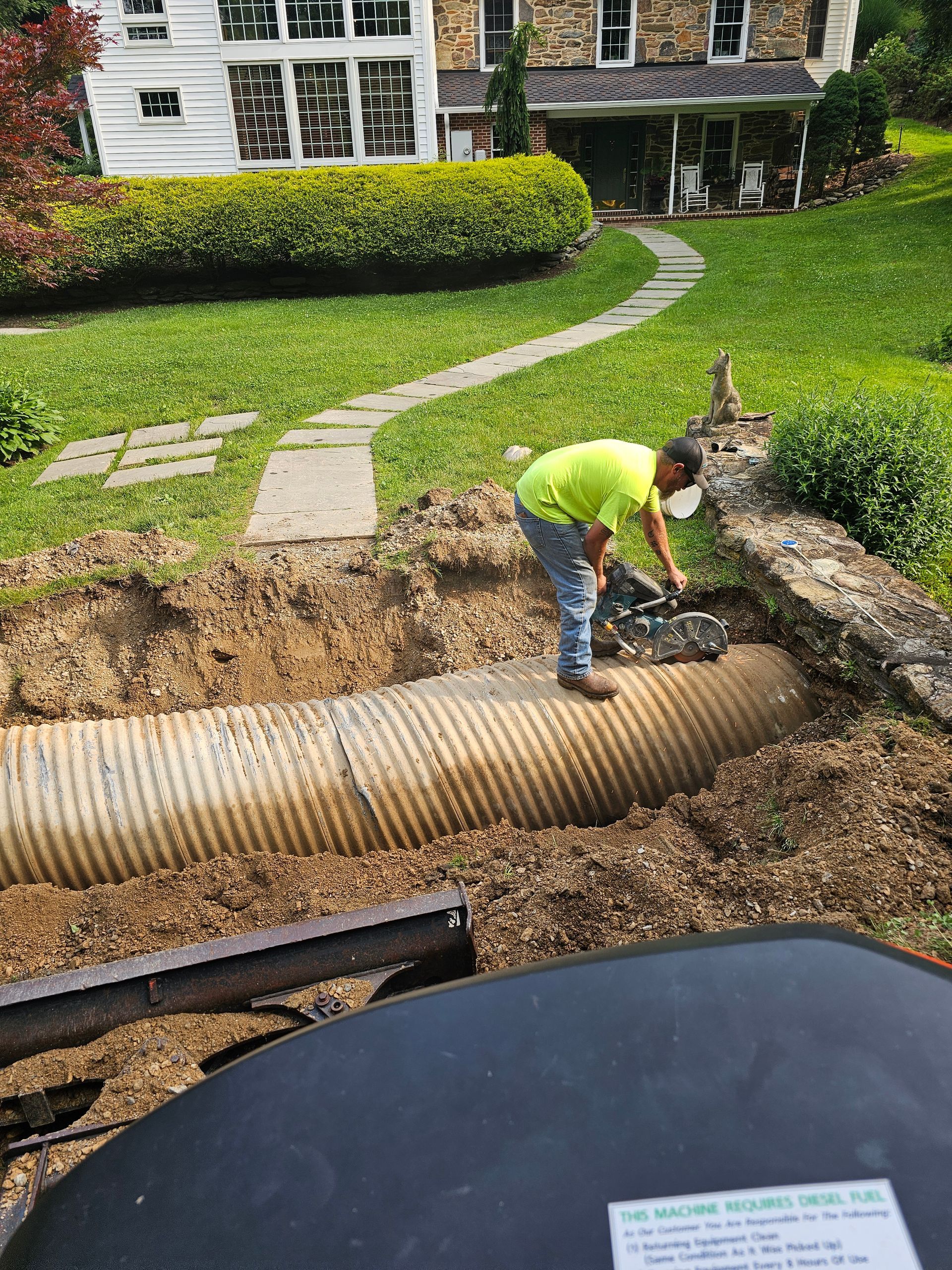 A worker in neon green shirt is working on a corrugated pipe in a dug-out area, with a house in the background.