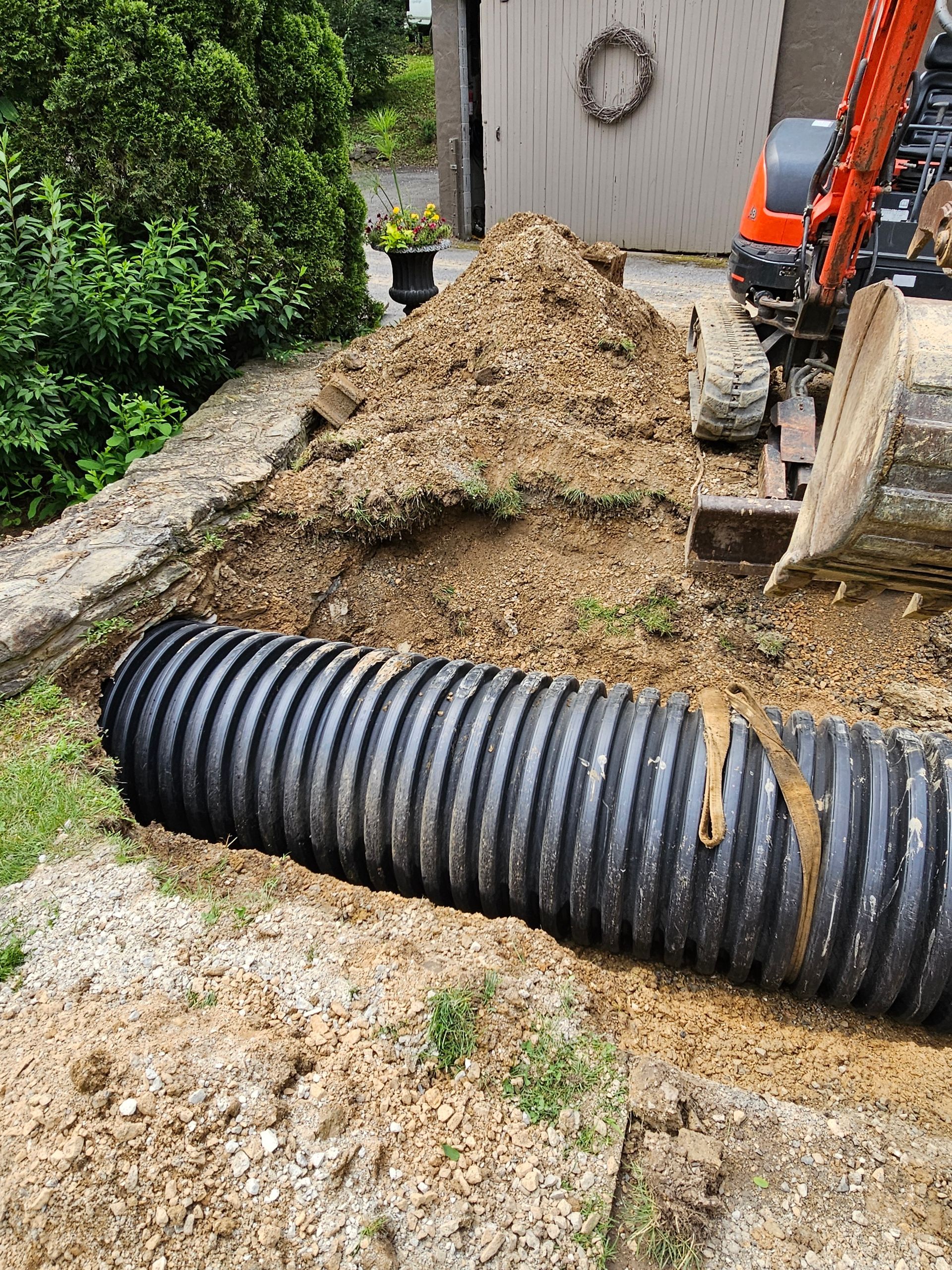 A black corrugated pipe in a trench with an excavator and dirt pile. Outdoors, daylight.