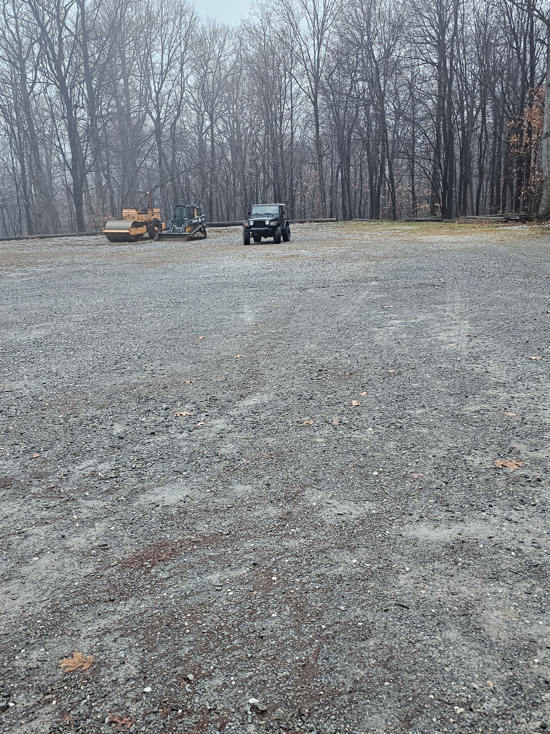 Gravel lot with vehicles: a black SUV, an excavator, and a motorcycle, with a blurred forest background.