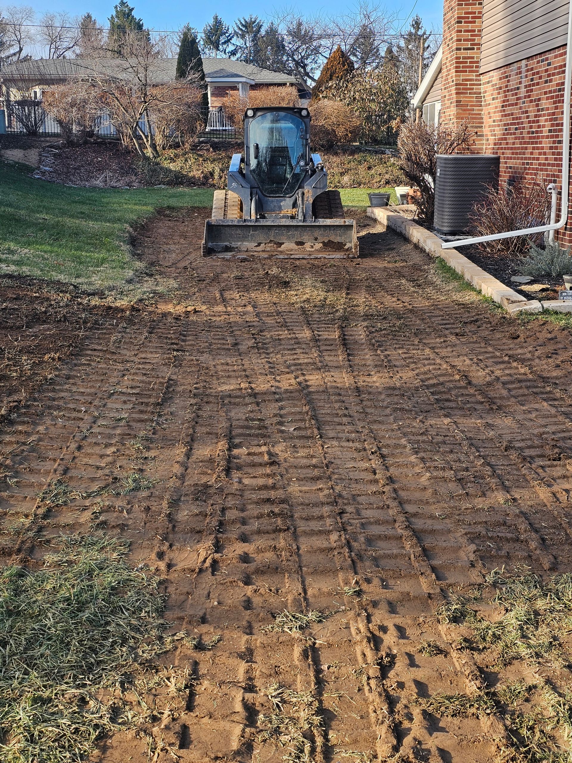Small bulldozer on a dirt area preparing ground. Brown soil, green grass, and a brick building are visible.