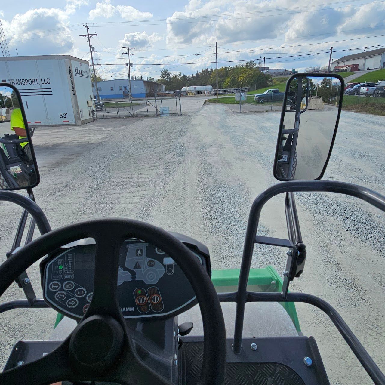 View from inside a machine driving on a gravel road toward a gate, with a worker visible on the left side.