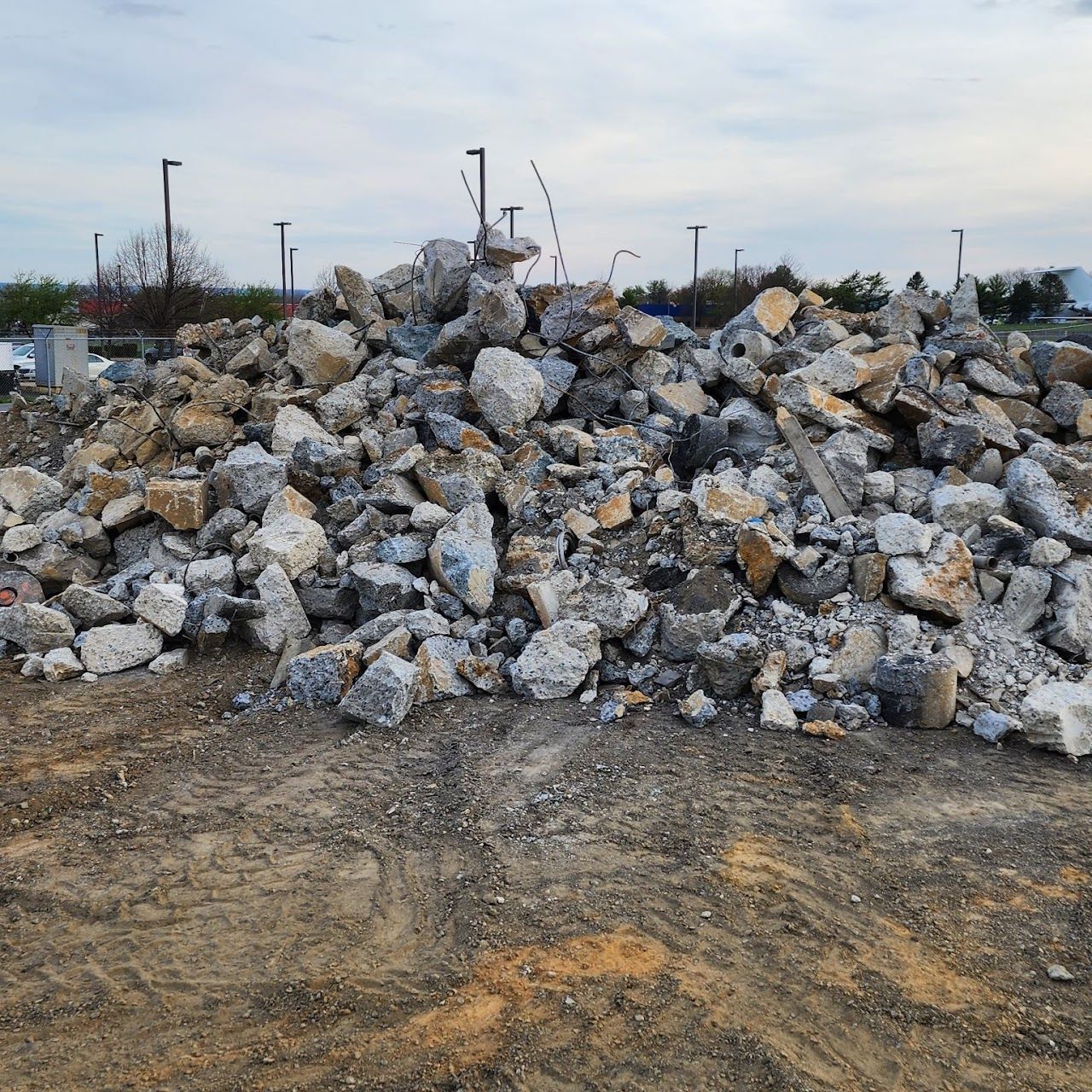 Pile of rubble, mostly grey concrete, sits on dirt. Background shows parking lot, cloudy sky.