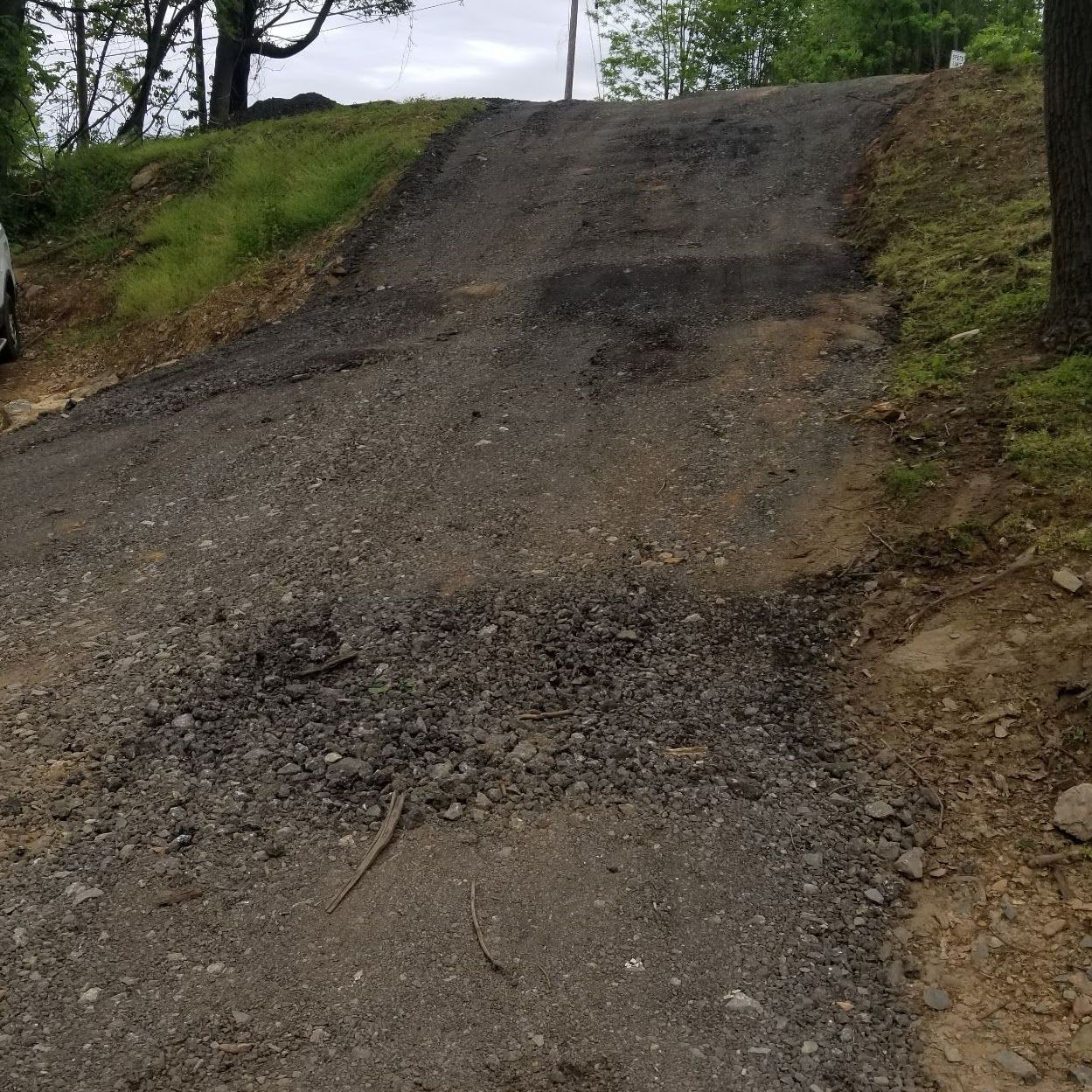 Gravel driveway leading uphill, worn asphalt surface, surrounded by grass and trees.
