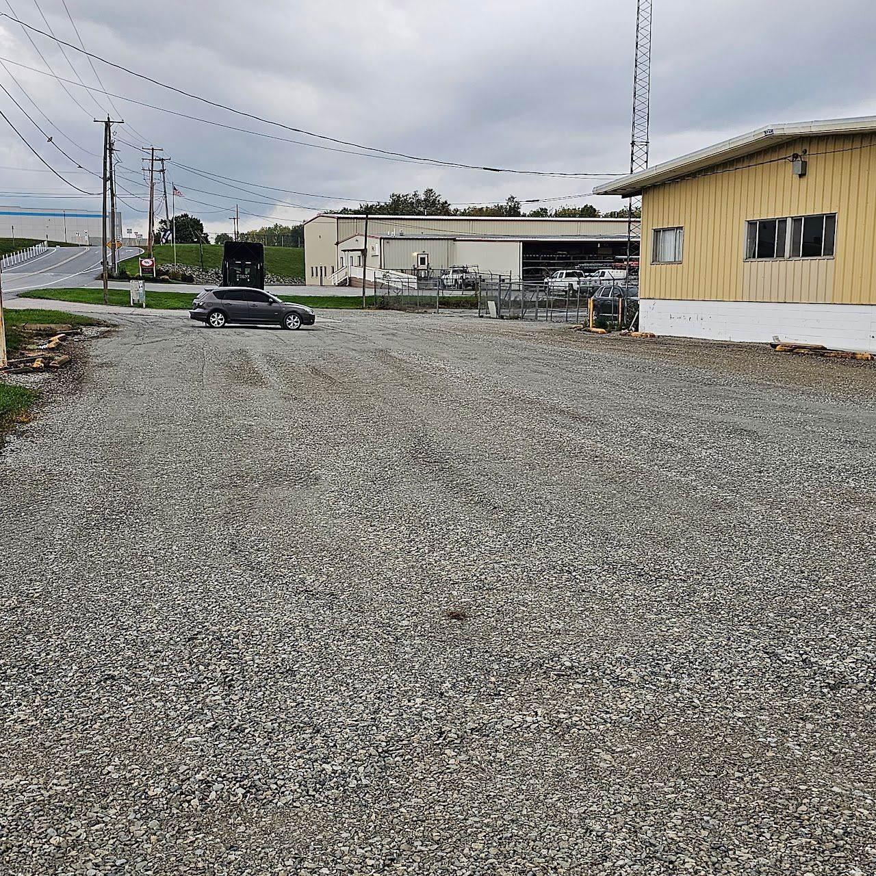 Gravel parking area next to a building and road, overcast sky. A dark car is in the parking area.