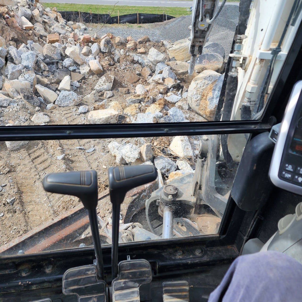 View from an excavator cab, looking at rocks and dirt. Levers and controls visible.