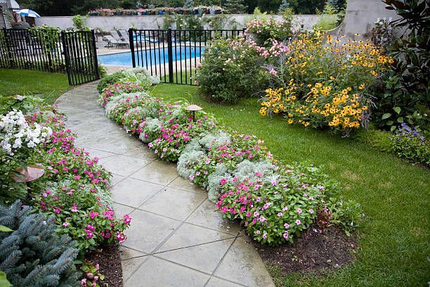 Stone path winds through a garden of colorful flowers and greenery leading to a pool behind a black fence