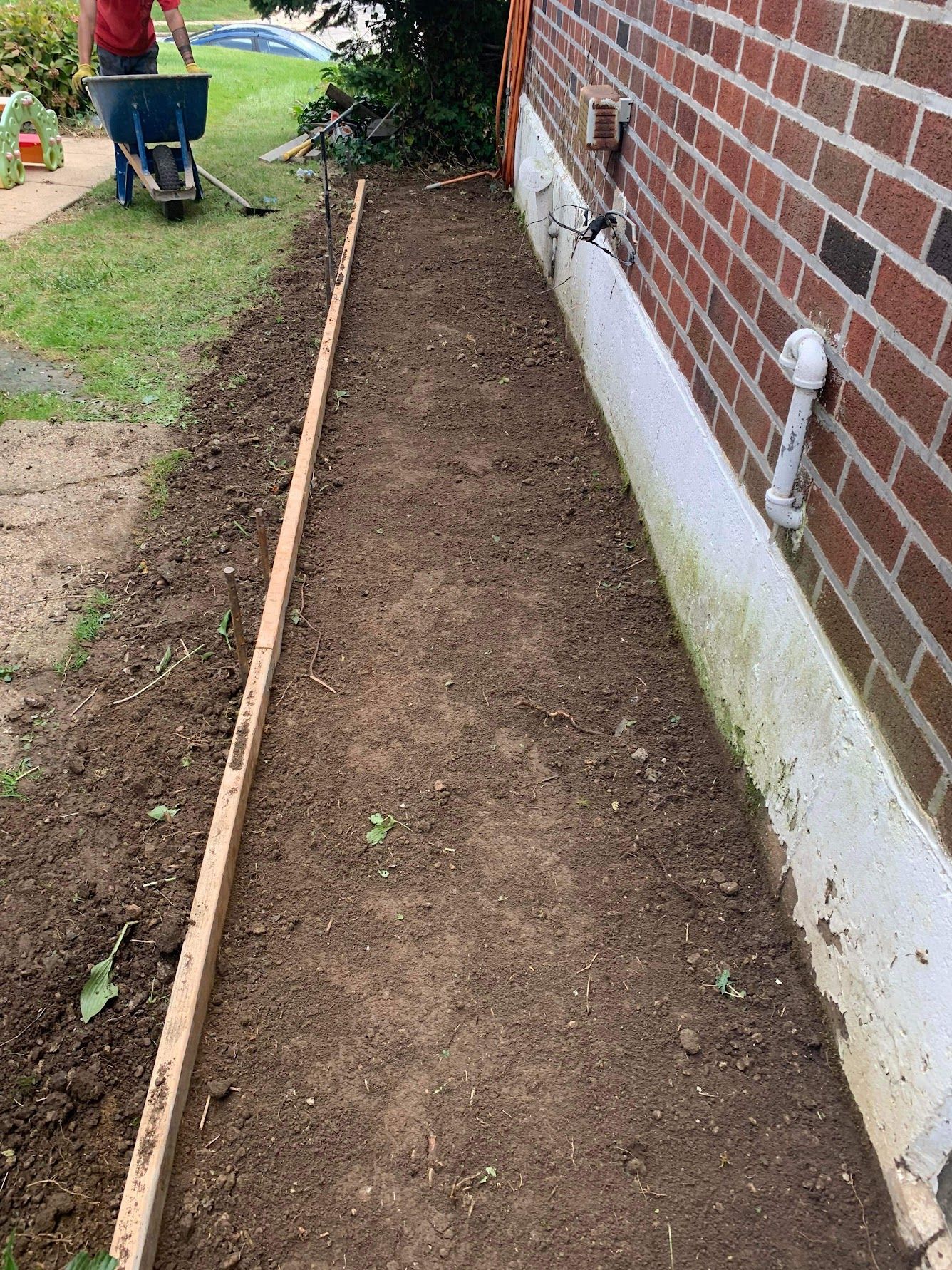 A person is pushing a wheelbarrow down a dirt path next to a brick wall.