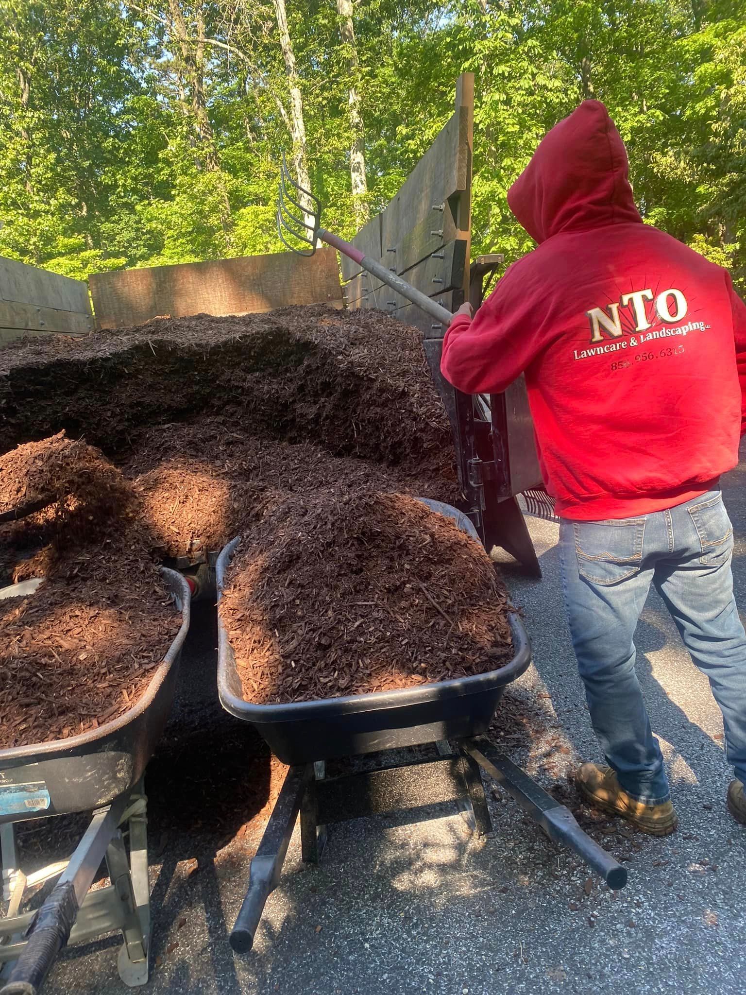 A man wearing a red nto hoodie is pushing a wheelbarrow full of mulch.