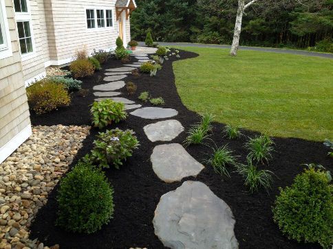 Stone pathway through a garden with dark mulch, bordered by shrubs and green grass next to a light-colored house installed by nto lawncare & landscaping in vineland, nj