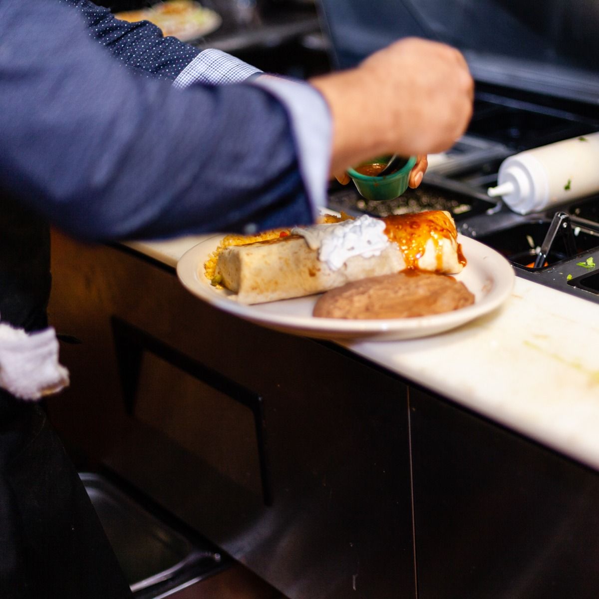 Chef plates food on a counter; sauces are added to the plate; kitchen setting.