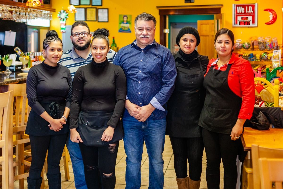 Restaurant staff posing. Six people stand inside. Warm lighting, yellow walls.