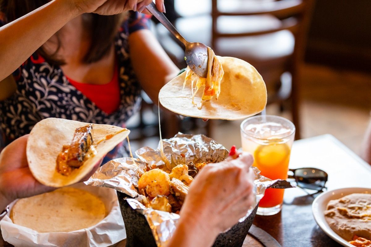 People scooping cheese from a bowl of food onto tortillas. A meal with drinks, inside.
