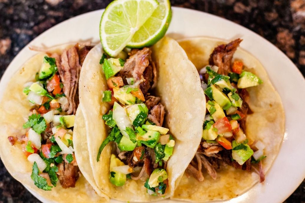 Three tacos with meat, avocado salsa, and lime wedge on a white plate.