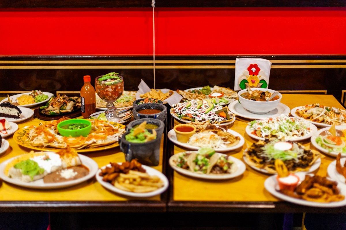 Table filled with various Mexican dishes: tacos, nachos, quesadillas, dips, salsa, and garnishes.