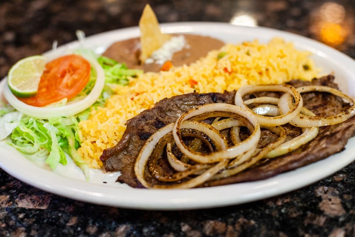 Plate of Mexican food: grilled steak with onions, rice, beans, lettuce, tomato, lime, and tortilla chip.
