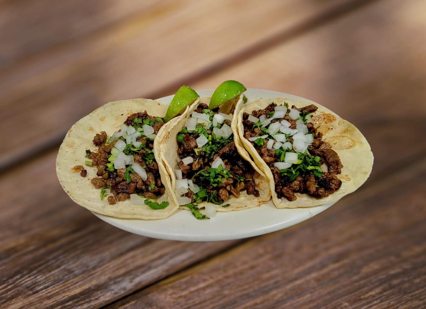 Three tacos on a white plate, filled with meat, onions, and cilantro, with lime wedges.
