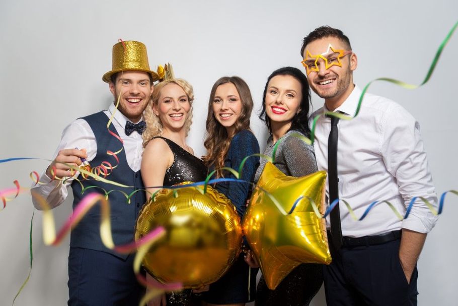 Four people at a party pose with gold balloons and confetti, smiling in festive dress.