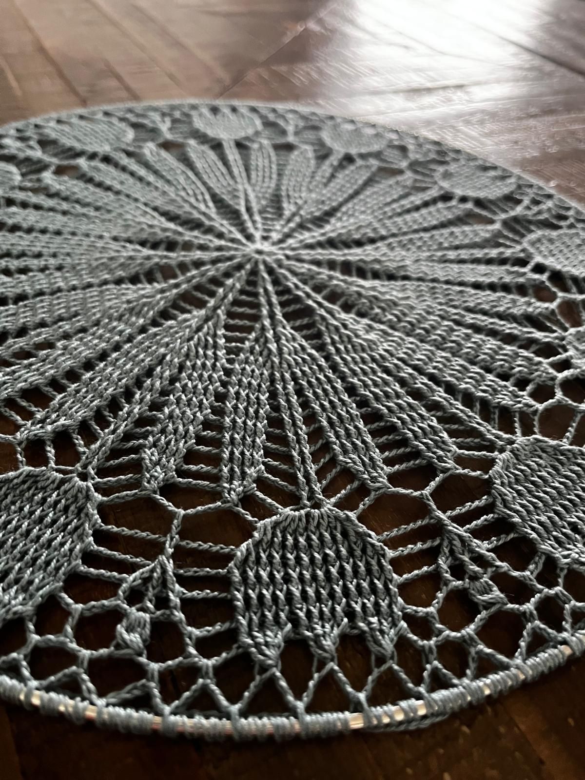 Een rond, grijs kleedje met een ingewikkeld kantpatroon, dat op een donkerhouten oppervlak rust.