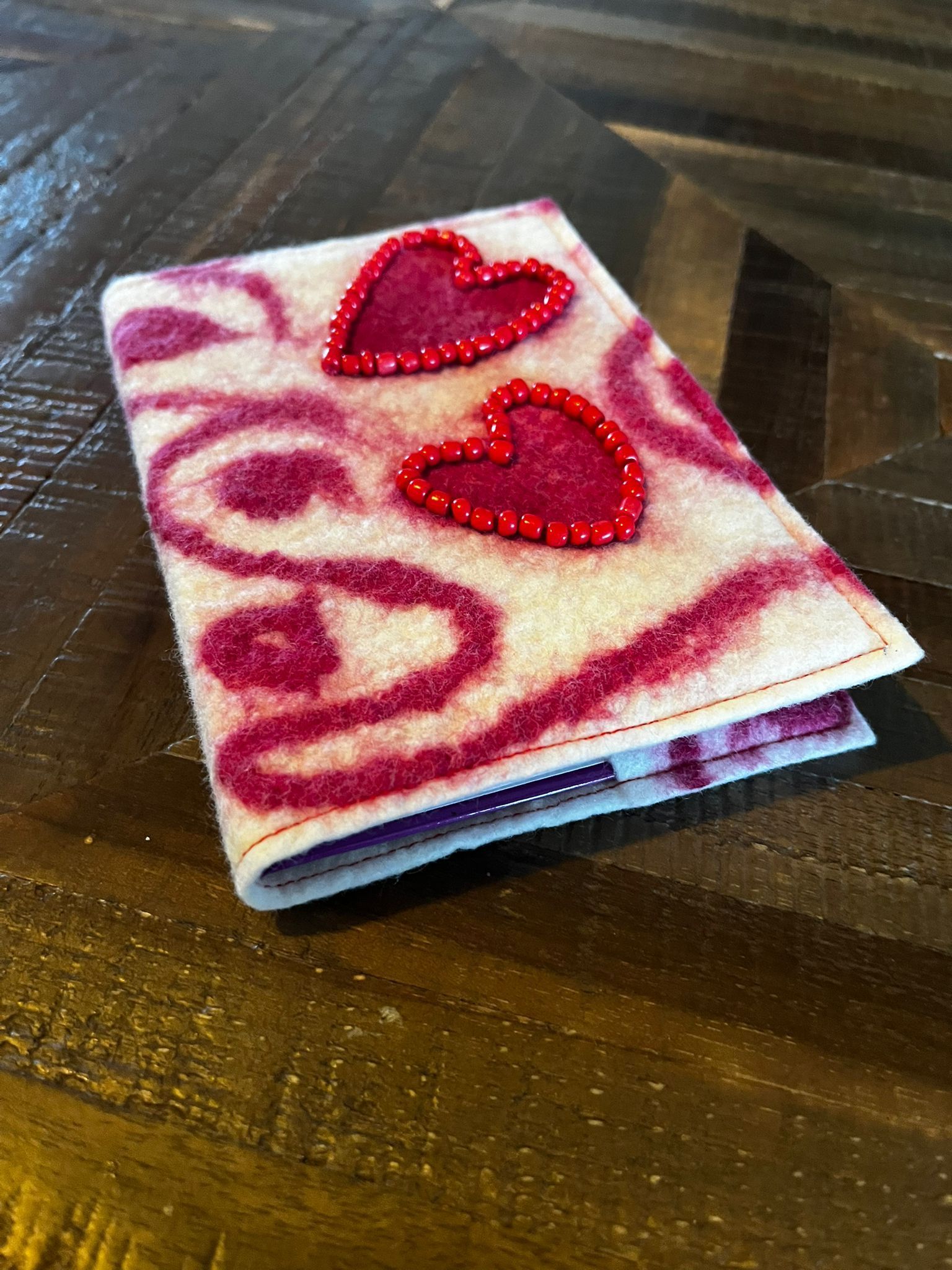 Een vilten portemonnee met twee rode kralenhartjes in een spiraalvormig patroon, die op een houten tafel ligt.
