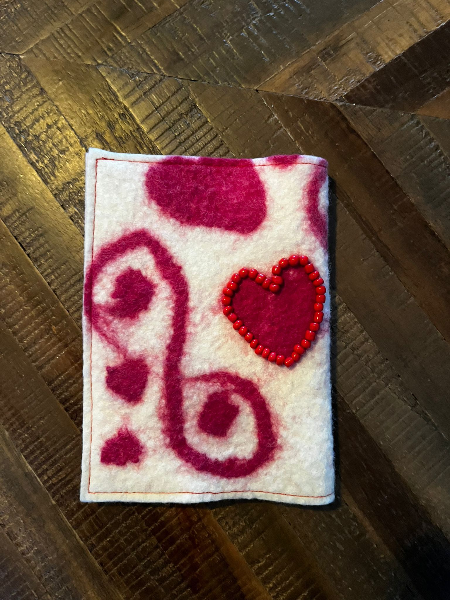 Een vilten portemonnee met twee rode kralenhartjes in een spiraalvormig patroon, die op een houten tafel ligt.