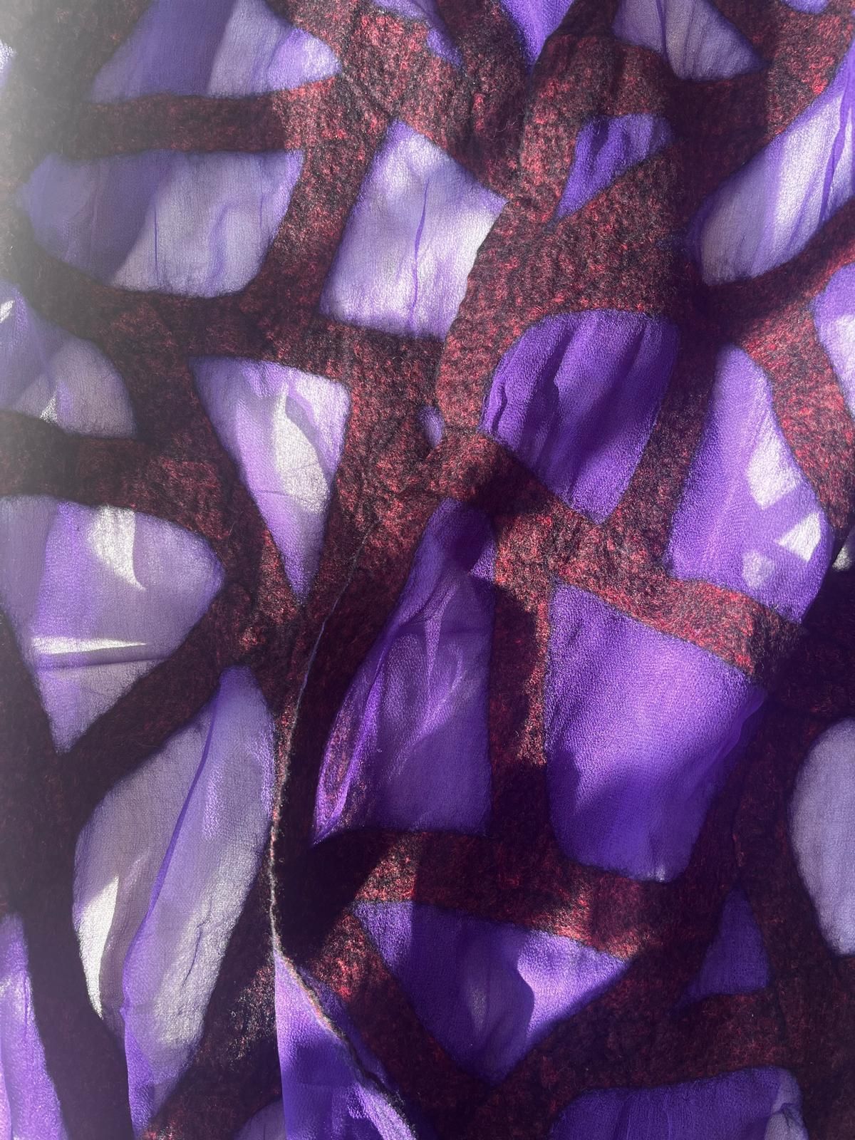 Paarse sjaal met donker geometrisch patroon, gedrapeerd over een wit kussen in het zonlicht.