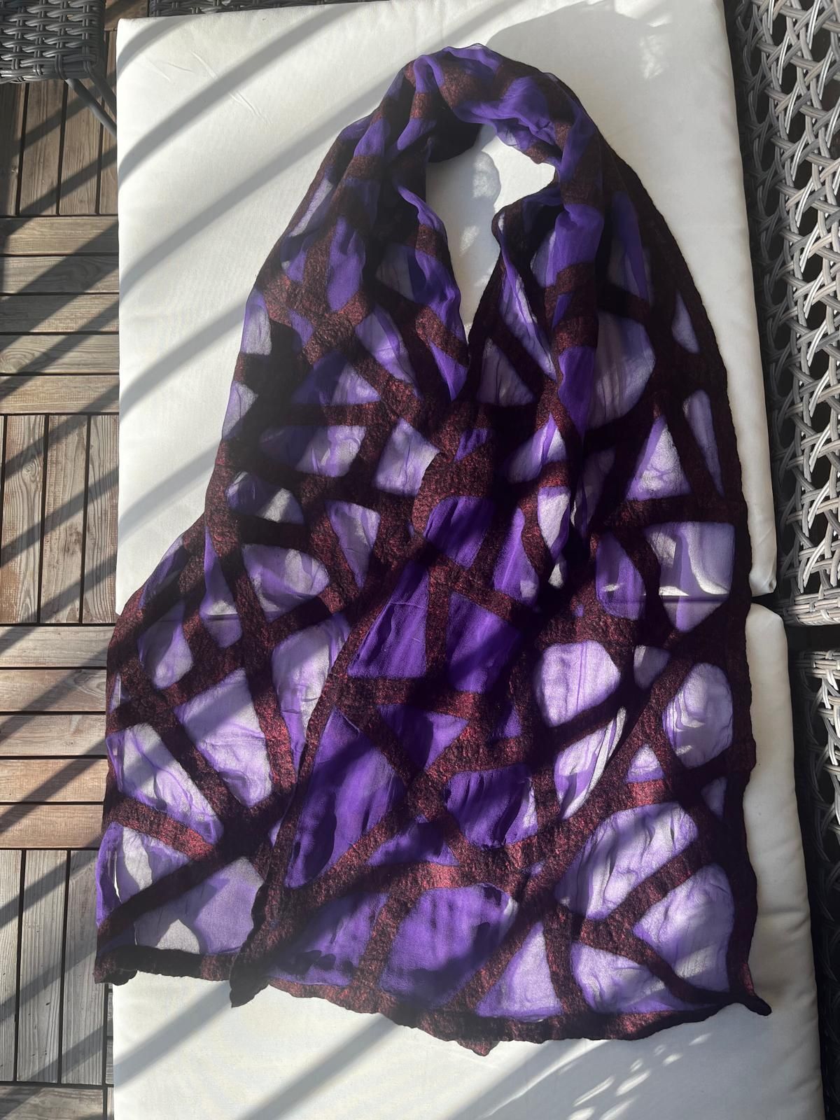 Paarse sjaal met donker geometrisch patroon, gedrapeerd over een wit kussen in het zonlicht.