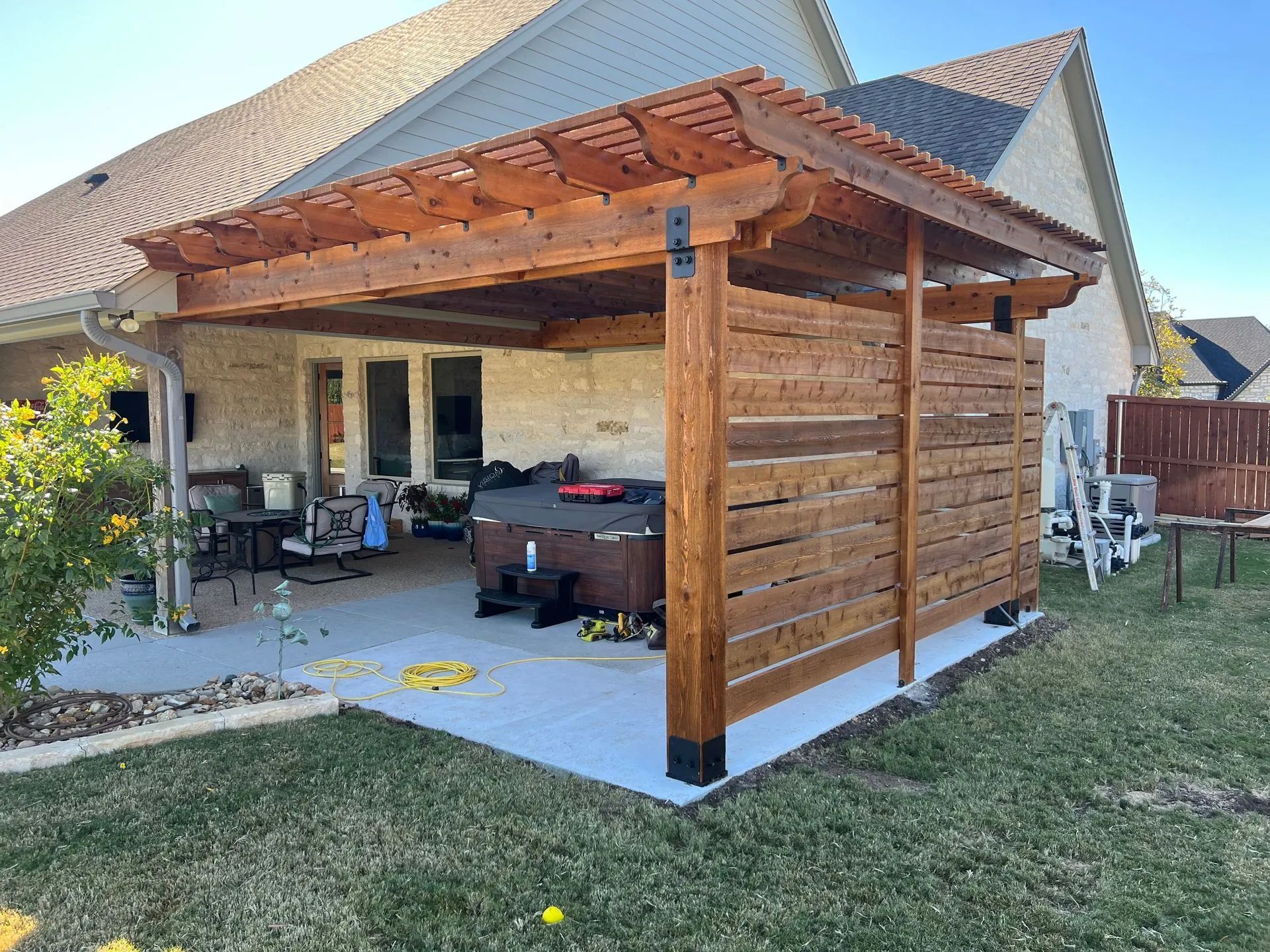 Wooden patio pergola with slatted roof, concrete floor, outdoor seating, and grassy yard beside a house