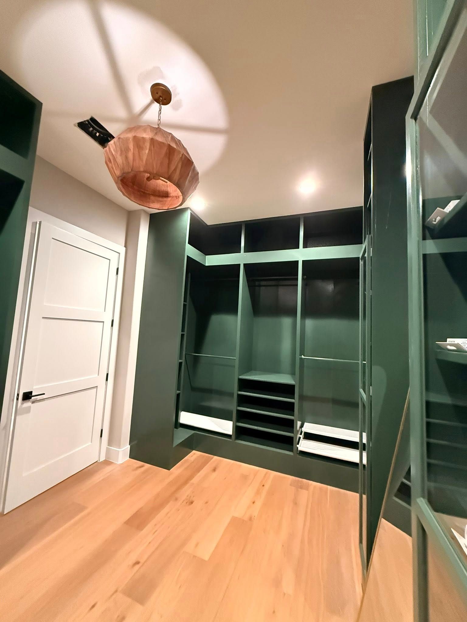Modern walk-in closet with dark green built-ins, white door, light wood floor, and woven pendant light