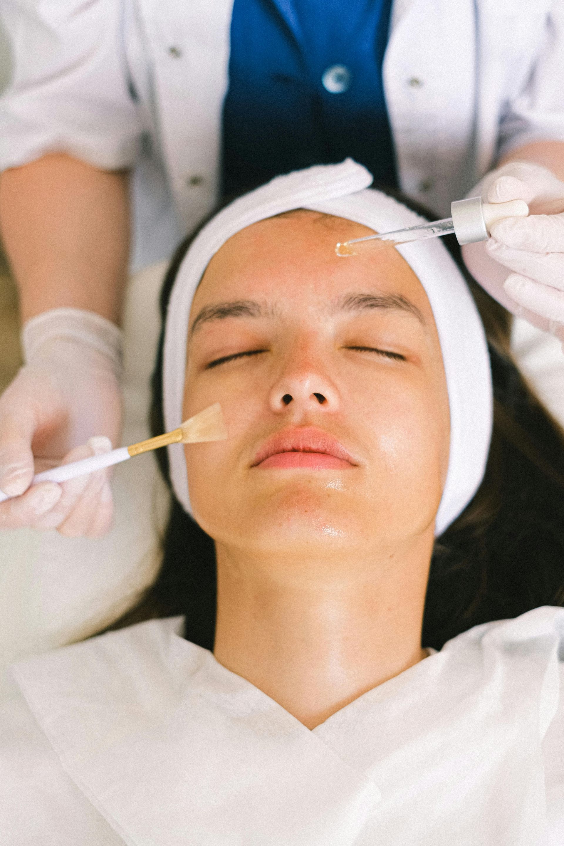 Woman receiving facial treatment, white headband, esthetician applying serum with pipette and brush.
