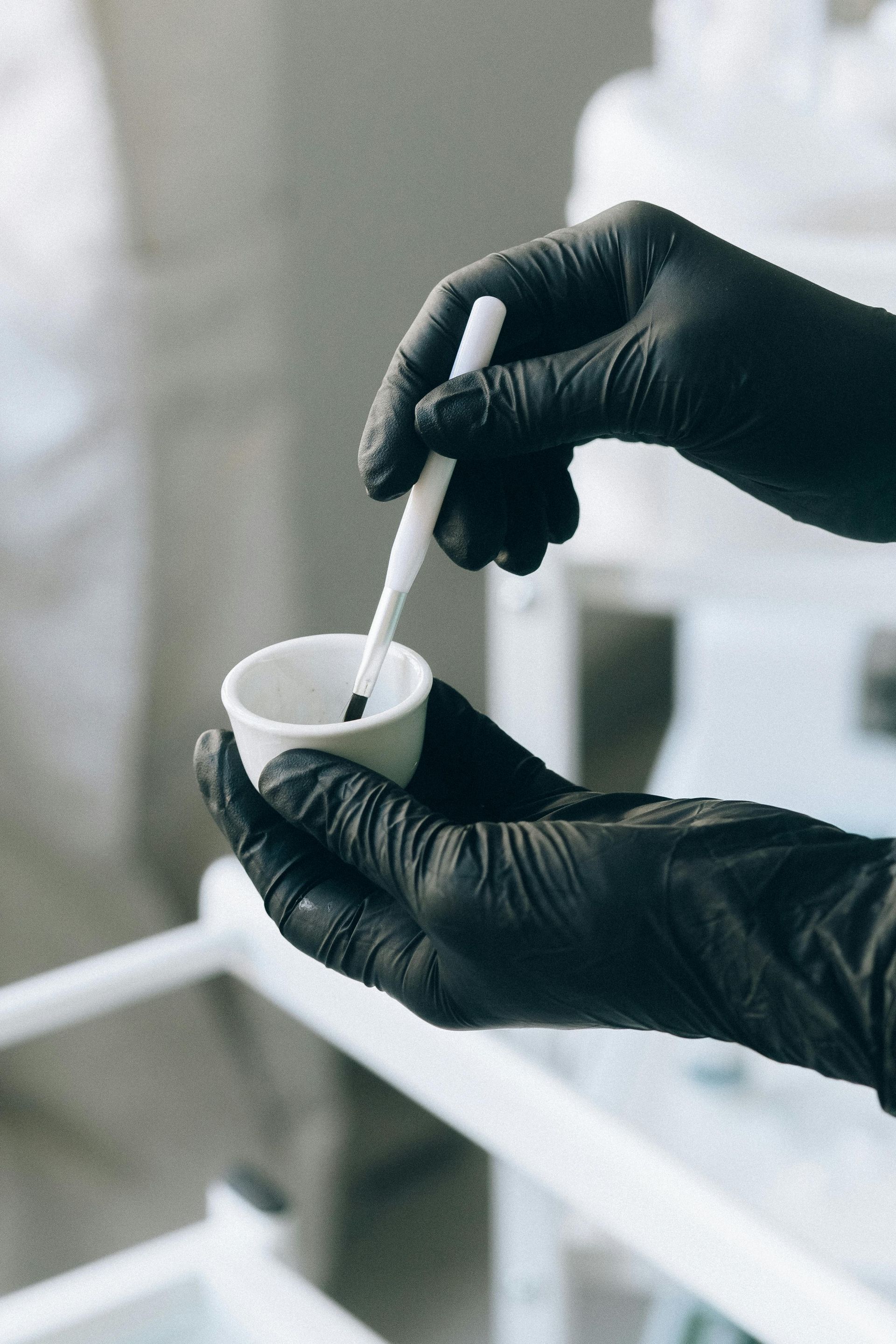 Hands in black gloves stir contents in a small white cup with a white-handled tool.
