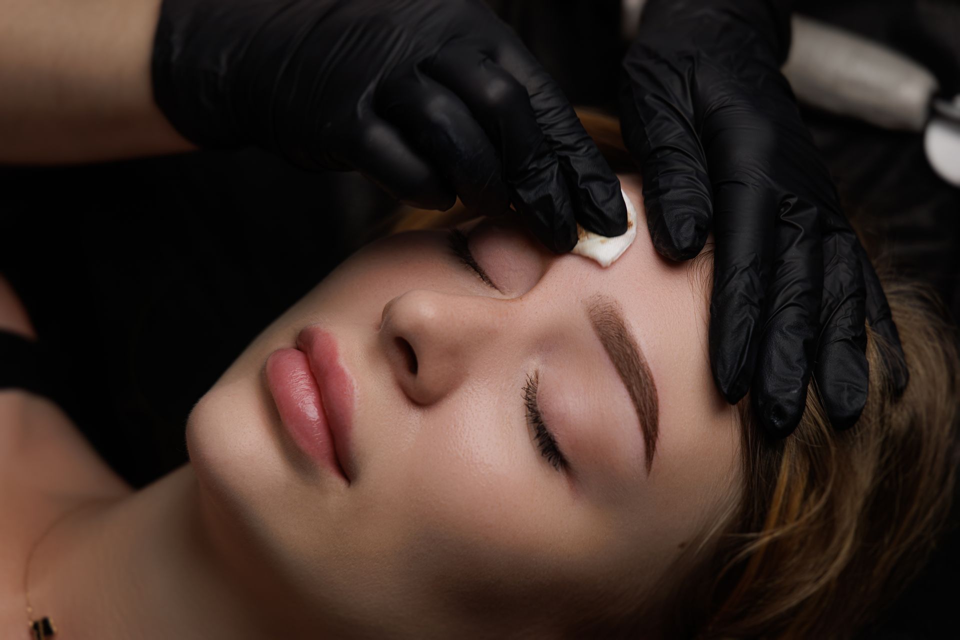 Person receiving eyebrow treatment, eyes closed, hands in black gloves applying cotton pad.