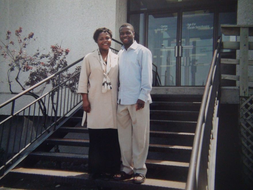 Man and woman smiling, standing on steps in front of building. Beige and blue outfits.