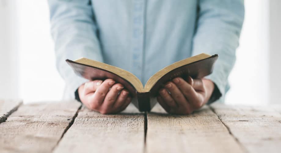 Person holding an open book with dark brown cover on a wooden table.