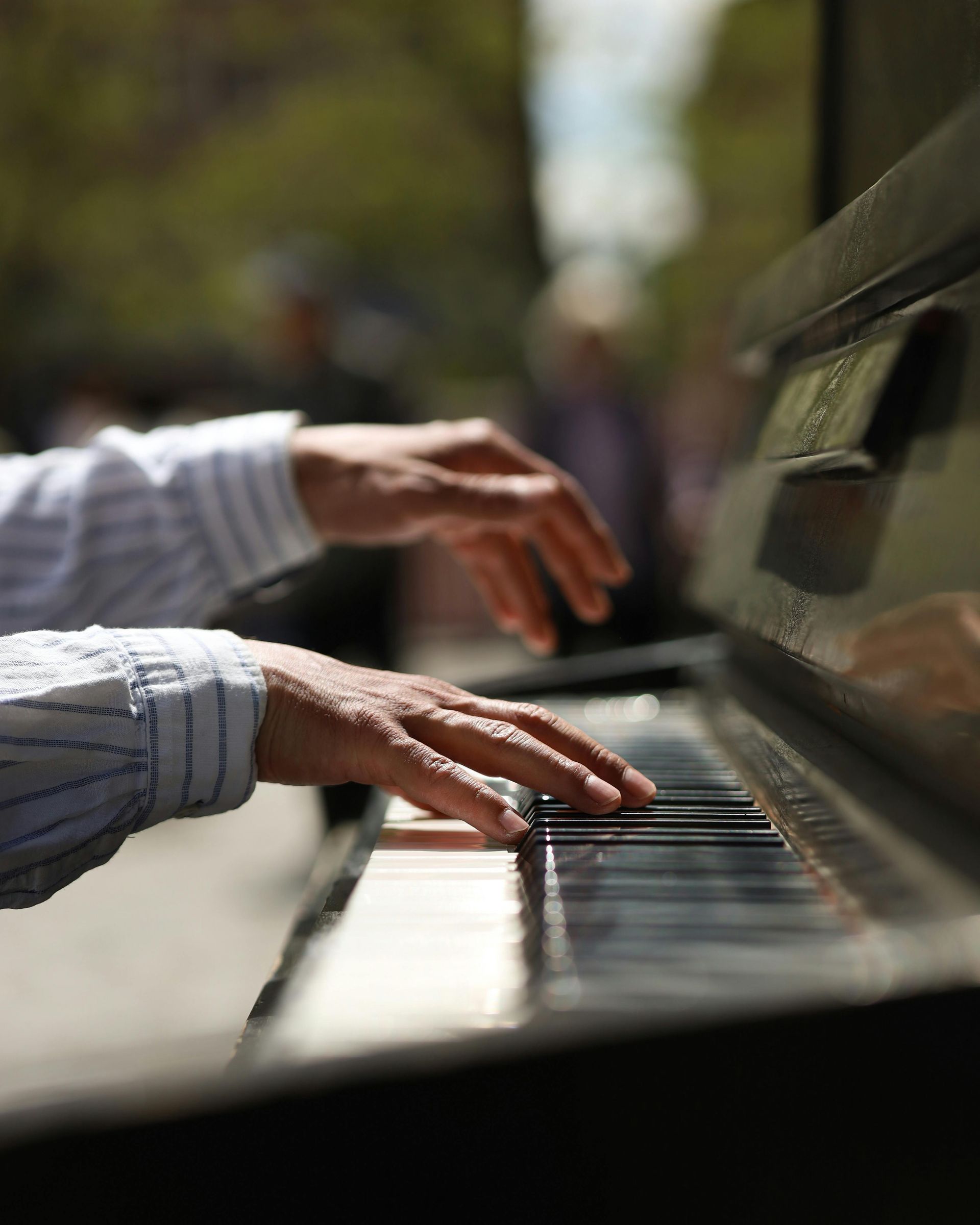 Hands playing a piano in an outdoor setting, wearing a striped blue and white shirt.