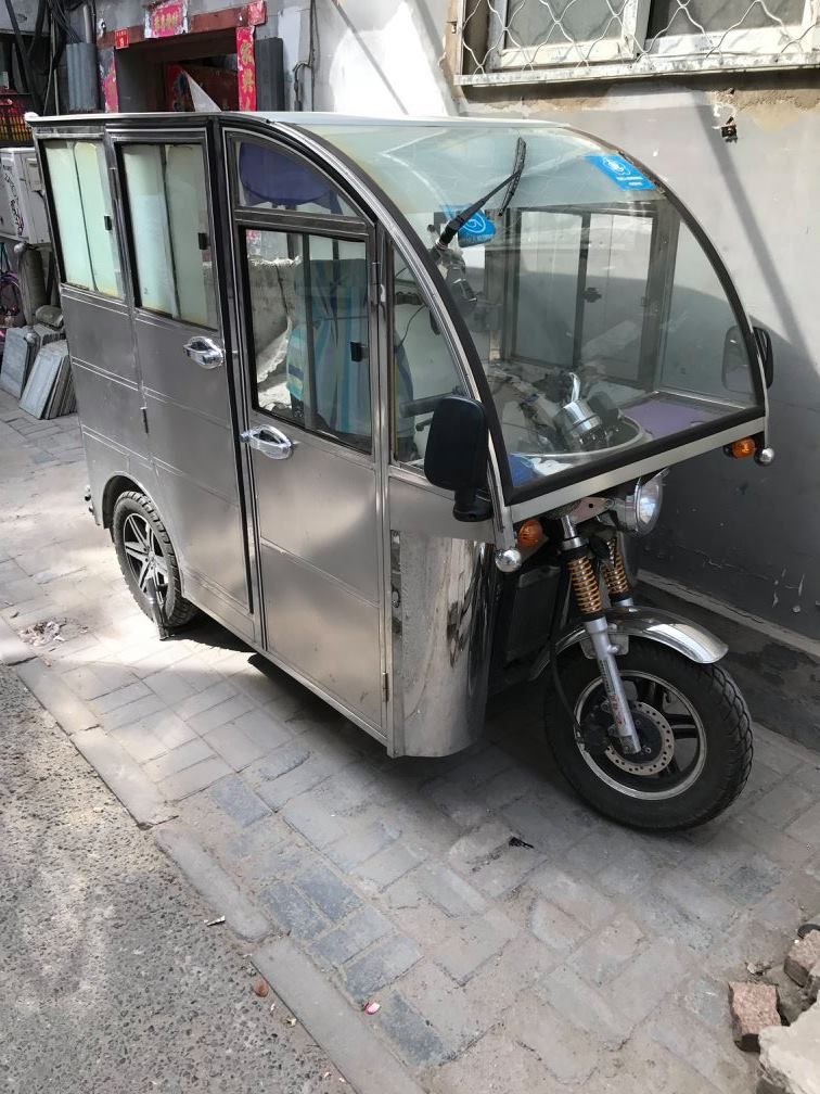 Silver enclosed three-wheeled vehicle parked on a brick-paved sidewalk.