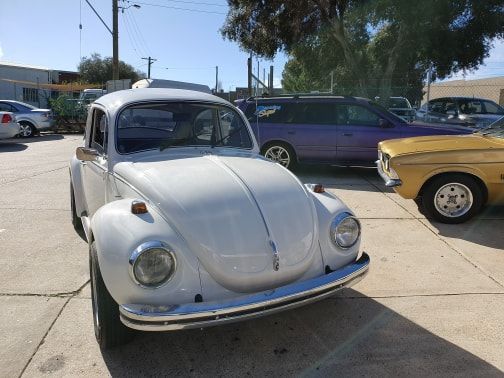 White Volkswagen Beetle parked in a lot, other cars visible in the background. Sunny day.