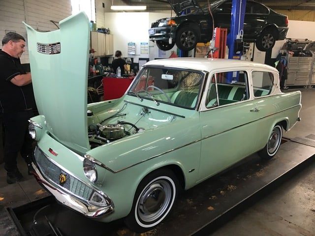 Pale green vintage car with open hood in a garage, a mechanic looks on.