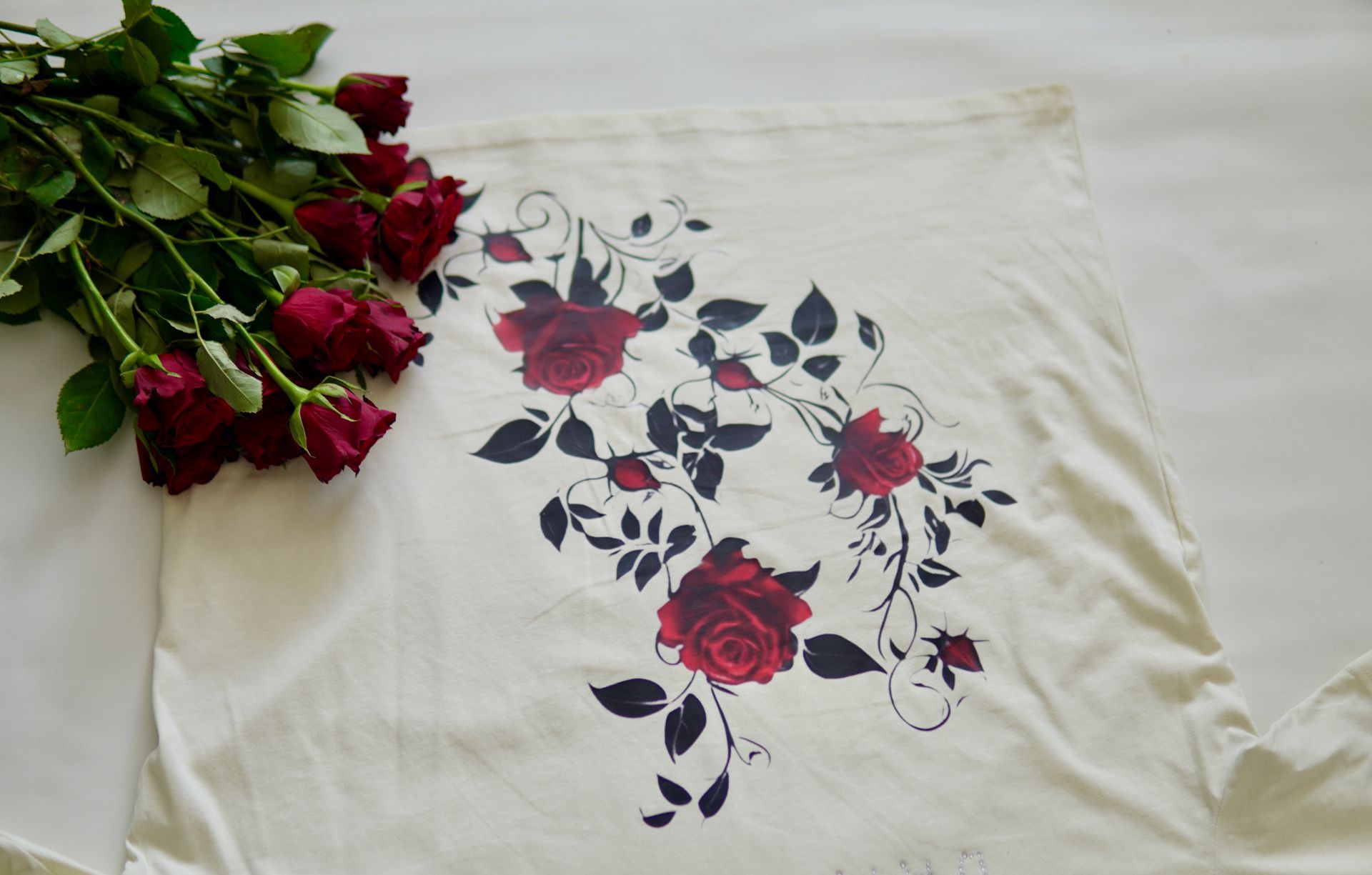 Cream-colored t-shirt with a floral design and a bouquet of red roses on a white surface.