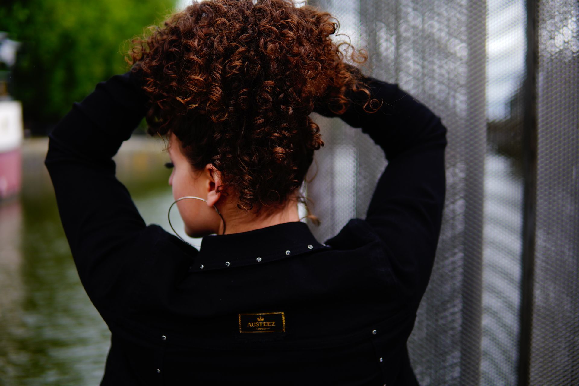 Person in a black coat adjusts curly hair outdoors near a chain-link fence.